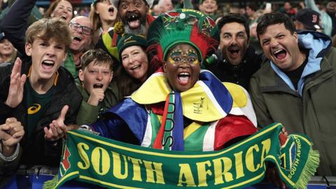 A female fan in brightly dressed South African costume stretches out a scarf that reads South Africa while other fans celebrate around her in the Stade de France after the Springboks win the 2023 Rugby World Cup final