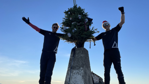 Ed Jackson and Ross Stirling at the top of Ben Nevis. They have their arms in the air and they are holding a decorated Christmas tree.
