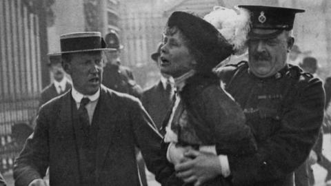 Policeman clasps Emmeline Pankhurst as another man walks along them