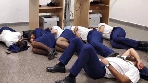 Six people dressed in airline uniforms lie on the floor of an office room