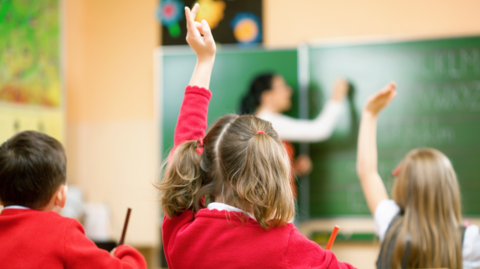 Primary school children are sitting at desks in their classroom. Their uniform is a red jumper. One girl is wearing a grey dress. A teacher is writing with chalk on the board. A girl with pigtails has her hand up.