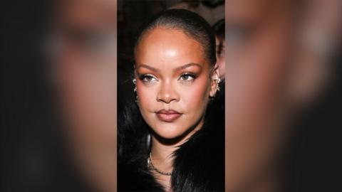 Woman with black hair tied back, ear piercings, and a black fur collar on her clothing.