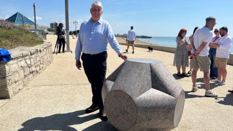 John Maine stands next to his latest sculpture, the sixth Portsmouth Marker. It is a large grey bolder carved in the shape of a hexagon and Mr Maine stands to the left of it. He is wearing a pale blue long sleeved collared shirt with black trousers and a pair of brown lace up shoes. He is standing on the promenade, there are crowds of people enjoying the sunshine in the background and the glass Pyramids building can be seen in the distance. The sea is also in vision as well as Southsea Pier.