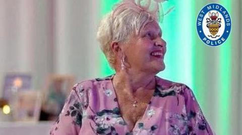 A woman in a pink floral style top, with a fascinator on top of her short blonde hair is looking to the right and smiling. There is a West Midlands Police crest at the top right of the picture