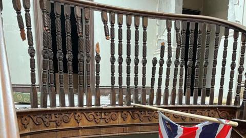 A broken brown ornate staircase with a union flag hanging. The walls are cream and there the ground is rubbly and white. 
