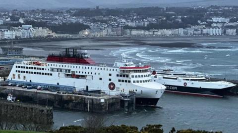 The Manxman and Manannan ferries moored in Douglas Harbour. The are both white with red and black trim. It is low tide in Douglas Bay and Douglas seafront is in the background.