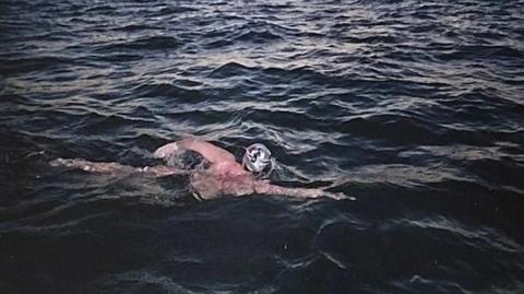 Dr Chrissie Thirlwell swimming in open water. You can rather dark and almost moody grey looking water with small triangular-shaped points as it slightly moves around her. 
Dr Thirlwell wears a silver hat and you can see her back with a black swimming costume. Her right arm is stretched out in front and her left elbow is raising out of the water as she turns her head to the left to breath in a front crawl stroke. Her legs are near the top of the surface in a kicking stance. Water fills the picture around her.