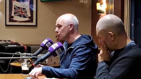 Two men sat a table with microphones inside a pub. Both men are bald and are wearing navy blue jumpers. 