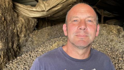 Lynden Croft is wearing a blue T-shirt with 'polo jeans' written on it in a logo. Behind him is a heap of 70 tonnes of potatoes in a big barn.