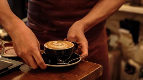 A barista is holding a cup with a cappuccino in it. A leaf pattern is on the top of the cappuccino. A teaspoon has been placed on the side on a saucer. 