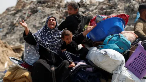 A woman, with her arms stretched out, sits on top of a wagon carrying her belongings and young children through rubble 