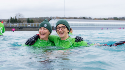 Two women in water wearing green swim rash vests and green woolly hats. They are both smiling and have their arms around each other.