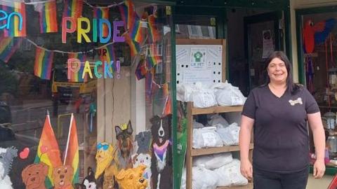 A woman in a black v-neck t-shirt with a bone shaped logo on the upper left side stands in front of a pet shop. To the left the shop window is decorated with pictures of dogs and rainbow lettering that says "Pride pack!". Behind the glass pride flags are visible hanging in the window