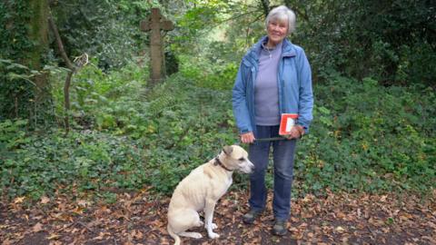 A woman with grey hair wearing a blue jacket and blue jeans stands with a light colured dog in an overgrown graveyard