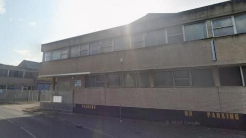The former Welton Bibby & Baron site. A two-storey building casts a shadow over the road. To the left of the building is an entrance to the site with a metal gate, which is open. 