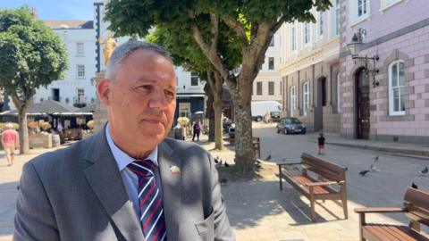 Constable Andy Jehan is stood in the Royal Square. He is wearing a grey blazer and a blue shirt.