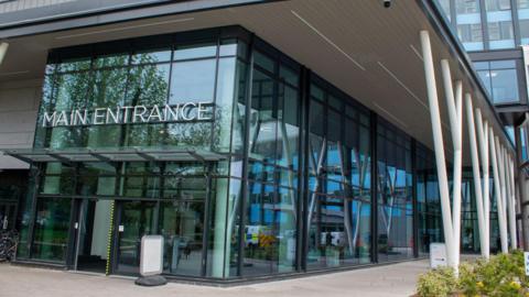 Large glass-fronted entrance to a modern building. There are white support beams and a sign which reads Main Entrance.