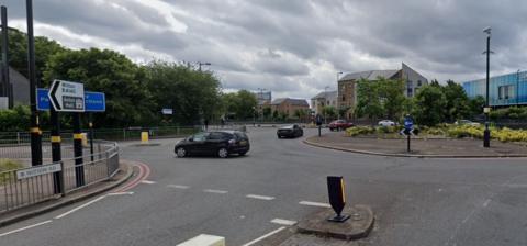 The approach to a large roundabout, with a sign showing Witton Road in the foreground and a road sign pointing to Witton and Aston Hall. The centre of the island has grass, low bushes and a tree growing. Four cars can be seen travelling around the island and mid-rise buildings can be seen in the background, including one clad in blue tiles.