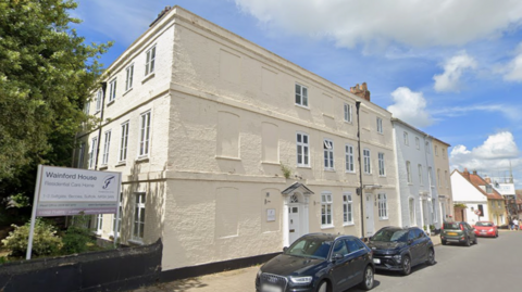 A general view of the exterior of Wainford House in Beccles. It is a three-storey building painted white. Some of the windows have been boarded up and painted over. Several cars are parked up outside the home.