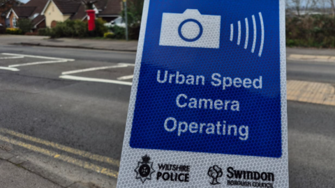 Image of a blue and white temporary road sign which shows an icon of a camera and the words: "Urban Speed Camera Operating" in the background can be seen an urban road, with housing and a red post box.
