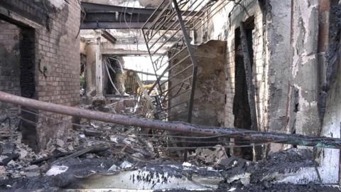 The inside of a home wrecked by fire with rubble on the floor, trees and burnt metal scattered around on the ground.