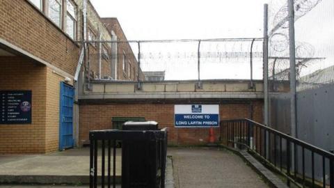 Part of a prison which has a sign saying "welcome to Long Lartin Prison". There are brown walls and a blue gate in the shot.