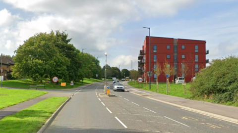 A google maps street view of Airport Road in Hengrove. The road is wide and there is an island for pedestrians in the middle. There are pavements either side of the road, grass verges and trees. There are cars travelling on the road and 30mph road signs on the roadside.