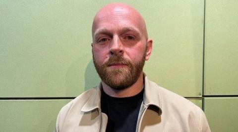 Sean Egan is looking at the camera, in front of a green wall. He is bald and has a beard, and is wearing a light-beige jacket with a black T-shirt.