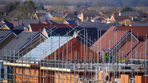 Scaffolding surrounds new build houses on construction site, there are two workers in hi-vis yellow jackets on one of the building's roof.