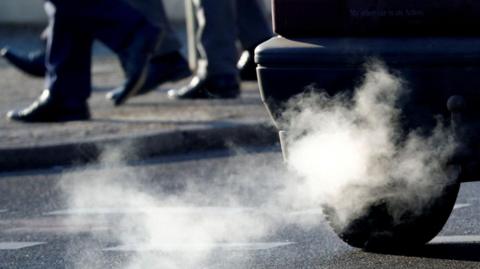 The rear of a car is visible with fumes coming out of the exhaust. Pedestrians are visible in the background. 