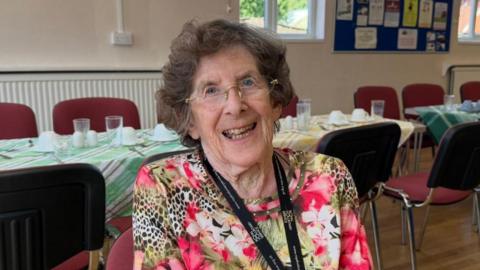 The photo shows Jennifer Hodge smiling at the camera. She has brown hair and is wearing glasses. She is also wearing a leopard print and flowered top, with a black lanyard around her neck. She is sitting in front of a table and chairs. The table has been set for tea.