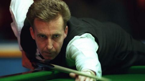 Terry Griffiths bent over the snooker table, cue resting on the bridge between his thumb and index finger on his right hand as he prepares to strike the white ball. He is wearing snooker attire, with a white long sleeve shirt and black vest.