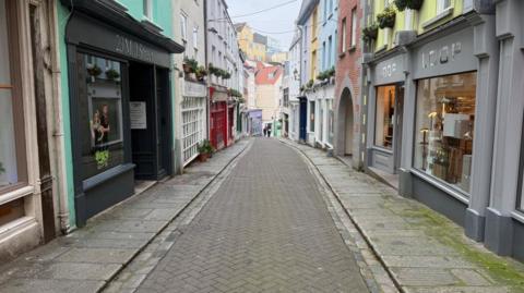 A shot of Mills Street. There are shops either side of a cobblestone road and two people walking side by side in the distance.  