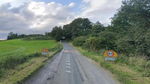 A country lane with road signs reading Elkstone on a summer's evening, with a large, open field to the left and bushes lining it on the right. It leads up a hill, and is covered by trees near the top.