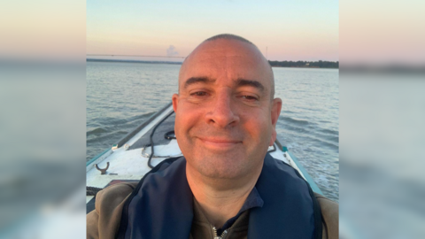 A man stood on a boat taking a selfie of himself. The sea is behind him and it is sunset.