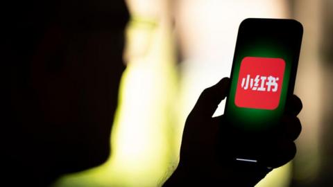 Backview of a man holding a phone, with the Xiaohongshu logo displayed on the phone.

