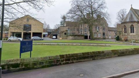A sign which reads REPTON SCHOOL with a number of stone buildings in the background