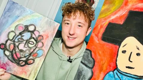 Alex holding two of his recent paintings. He is wearing a green hoodie and has curly brown hair. On the left is a painting of a face with circles around the edge and, on the right, is a picture of a man wearing a black hat.