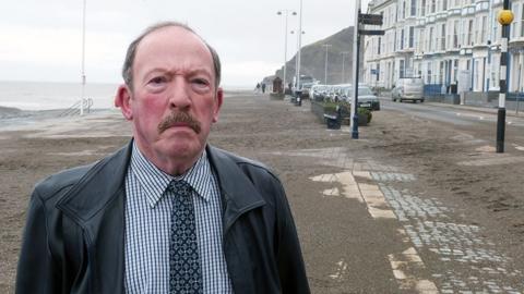 A picture of Alun Lloyd Jones looking into the camera. He's stood on Aberystwyth beach. 