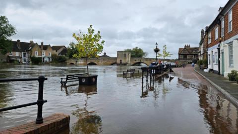 River flooding a path and covering benches and plant pots. Bridge in distance with archways nearly covered by the river showing how high it is