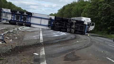 A white and blue lorry lies on its side across the central reservation with all the wheels and tyres showing and dirt, dust and white paint marks across the road.