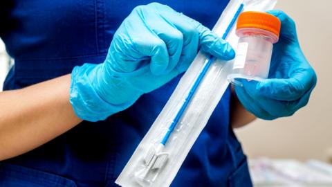 A person wearing navy scrubs and light blue rubber gloves holds a medical swab and container.