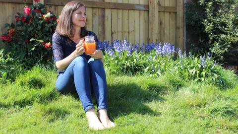 Maria sitting in a garden on the grass holding a cup of juice looking into the distance