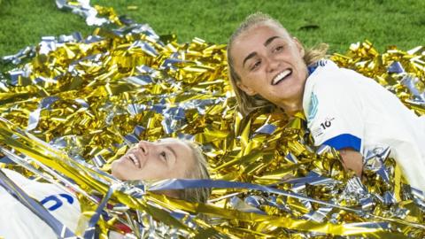 England's Leah Williamson and Georgia Stanway lie in gold and silver  confetti as they celebrate after winning the UEFA Women's Euro 2025 final match between England and Spain, in Basel, Switzerland. They both wear their blonde straight hair in a ponytail and are smiling.