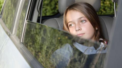 The image shows a child sat inside a car, viewed through the open window on the left side. The child has long straight, light brown hair and wears a school uniform with a white polo shirt and a grey pinafore. The car interior is grey, and greenery outside is reflected on the window.