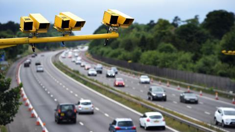 Yellow ANPR cameras in the foreground of the photo, below them is a motorway with cars on it, with trees in the background