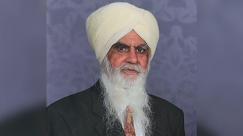 Bhai Joginder Singh wearing a dark suit, orange tie and a cream-coloured turban. He has a long white beard.