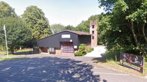 A Google Maps image of Mayfield Community Fire Station. It is brick building with a black, wooden feature. There are trees in the background. 