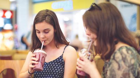 Two girls drink milkshakes in a cafe with a wooden table. 