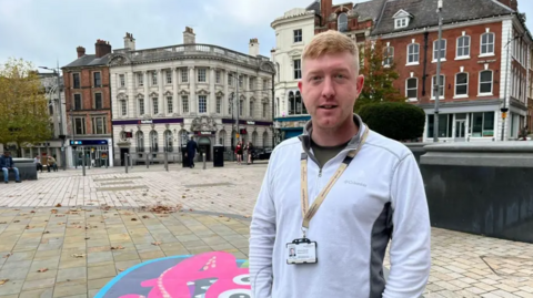 Simon is stood in a city centre square with three storey buildings behind him. He is wearing a white and grey top and a lanyard with an ID card in and has his hands in his pockets. He has short reddish hair.